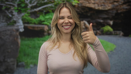 Blonde woman smiling broadly shows a thumb up gesture with her right hand in a sunlit park;...