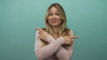 Blonde woman pointing with crossed wrists and index fingers in teal studio setting; indecision...