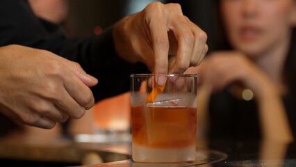 Bartender squeezing orange peel smelling over glass cocktail whiskey putting into Old fashioned on top of rock ice making special drink advertisement alcohol on Paloma and guest background. Vinosity.