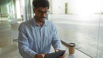 Young man with beard in clinic room holding tablet while seated at indoor table surrounded by modern interior design