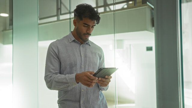 Young hispanic man in clinic setting reading digital tablet, wearing striped shirt and focused on screen, standing in modern, bright, indoor room environment with glass walls.