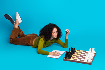 Young woman writing notes while playing chess on a vibrant blue background showcasing leisure and...