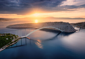 Aerial view of bridge and blue sea at colorful sunset. Modern Krk bridge island during golden hour, mountains, rocks, reflection in water and orange sky. Top drone view of bridges. Architecture