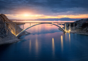 Aerial view of bridge and blue sea at night. Modern Krk bridge with illumination, island, mountains, water and cloudy sky at sunset. Top drone view of road, city lights, rocks at dusk. Architecture