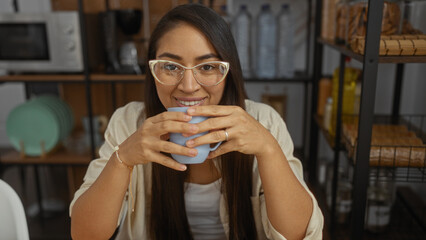 Young woman smiling with glasses holding a mug in cozy indoor apartment setting, showcasing a warm latin interior with a focus on home comfort and relaxation.
