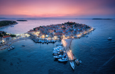 Aeriel view of old town with street lights on island at night. Summer in Primosten, Croatia, Adriatic Sea. Top view of houses, pink sky, water, city lights, boat, yachts, illuminated waterfront