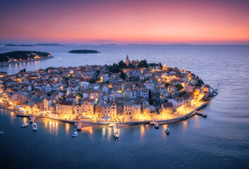 Aeriel view of old town with street lights on island at night. Summer in Primosten, Croatia, Adriatic Sea. Top view of houses, pink sky, water, city lights, boat, yachts, illuminated waterfront