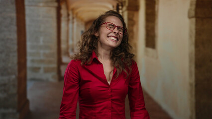 Fototapeta premium Woman smiling in red shirt with curly hair and glasses standing indoors at old university hallway.
