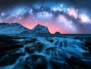 Naklejka premium Night starry sky with glowing Milky Way, rocky shore with stones in water. Winter colorful landscape with milky way and stars, fjord, pink sky, sea, snowy mountains in Lofoten Islands, Norway. Space
