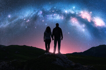 Couple of hikers holding hands standing on a rocky outcrop in mountains, night pink starry sky with glowing Milky Way. Colorful landscape with silhouettes of man and woman, bright stars and milky way