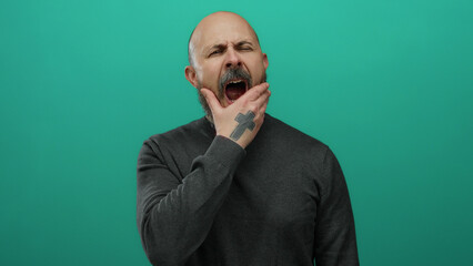 Man with a beard yawning against a solid green background, showcasing a bald head and grey sweater, exuding a handsome, tired expression in a minimalist setting.