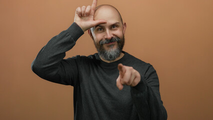 Man gesturing loser sign isolated against brown wall with bald head and beard wearing dark shirt in humorous expression pointing with finger