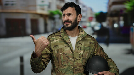 Fototapeta premium Hispanic man with beard in military uniform gestures outdoors on city street holding a helmet, showcasing urban scenario.