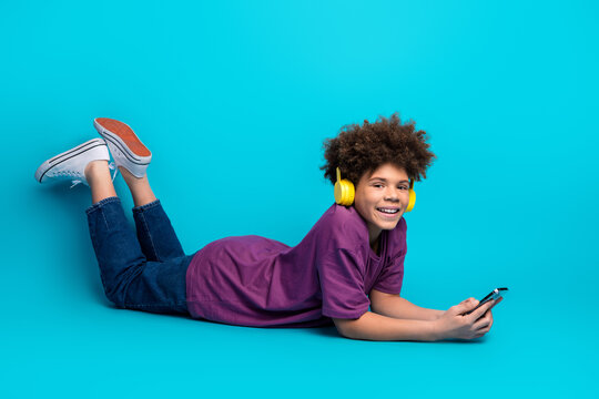 Smiling young boy listening to music with yellow headphones and using smartphone against vibrant blue background