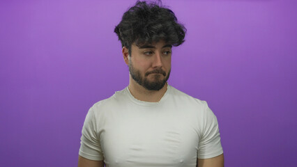Young hispanic man with beard in white shirt stands against vibrant purple background with...