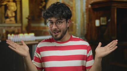 Young man with beard and glasses wearing striped shirt gesturing with surprise inside a church with...