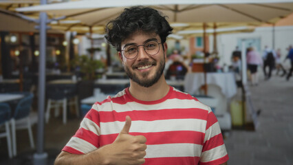 Young bearded man in glasses and striped shirt smiling and pointing to self with thumbs up gesture...