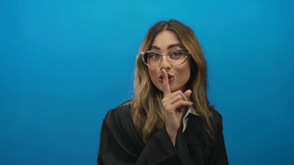 Female judge wearing glasses and judicial robe places finger to lips gesture in a studio with blue...