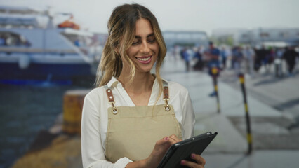 Blonde young woman wearing apron and white shirt taps tablet with fingers at pier; joy leisure...