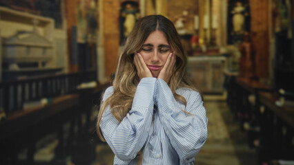 Young slender blonde woman holds bare hand on cheek in historic cathedral; toothache pain worry anxiety.