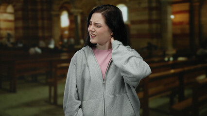 Woman experiencing neck pain while standing in a dimly lit catholic church interior with wooden...