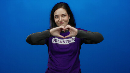 Naklejka premium Brunette woman in purple volunteer shirt gestures heart with hands against vibrant blue background, conveying emotion and dedication.