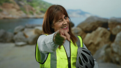 Hispanic senior woman winking and pointing at the camera in a vibrant outdoor setting at the beach,...