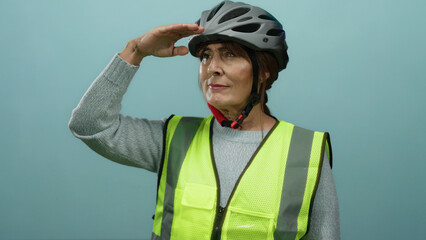 Senior hispanic woman with helmet and safety vest saluting while looking far with a hand on forehead against a blue isolated wall background.