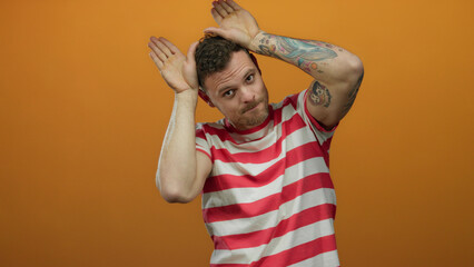 Young hispanic man in striped shirt makes playful ear gesture against isolated orange background,...