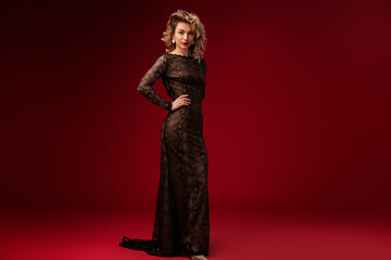 Chic blonde woman poses confidently in exquisite black lace gown against vivid red backdrop,...