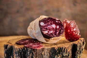 Dry-cured sausage in a fabric casing and sliced ​​pieces on a wooden background. Rustic atmosphere, natural products, home cooking. Ideal for illustrating traditional gastronomy.