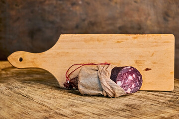 Sausage in a fabric casing lies horizontally against the background of a long cutting board. The concept of traditional gastronomy, natural products, home cooking, meat delicacies and snacks. 