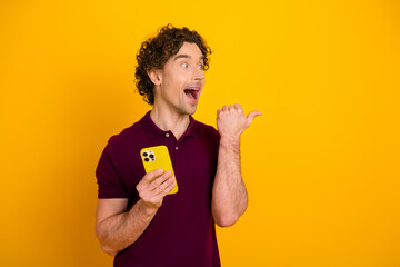 Excited young man in burgundy polo holding smartphone against a vibrant yellow background,...