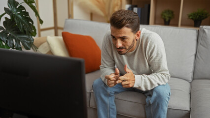 Young man relaxing in living room, watching television with focused expression, sitting on sofa, fingers interlocked, surrounded by cozy home decor, casual and stylish setting.