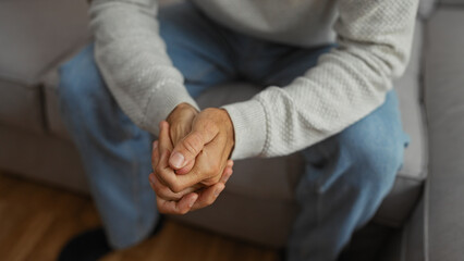 Young man sitting on a couch indoors holding hands together in a thoughtful pose wearing casual clothing in a cozy home setting