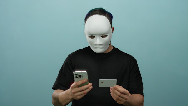 Young bearded man wearing a mask on a blue isolated background holds a smartphone and credit card, symbolizing anonymity and online interactions. - Powered by Adobe