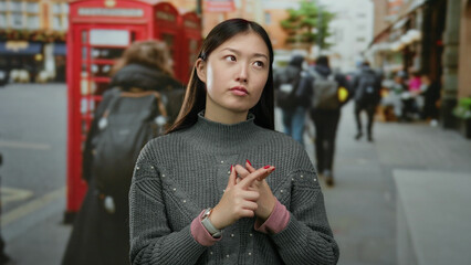 Woman standing outdoors near red phone box wearing gray sweater thoughtful expression urban setting...