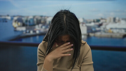 Woman outdoors by seaside at promenade appearing troubled with hands on face and blurred waterfront...