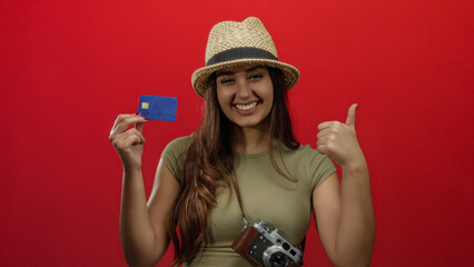 Woman smiling over isolated red background holding credit card and camera wearing casual hat...