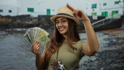 Fototapeta premium Hispanic woman at seaside holding peruvian soles, smiling and saluting with hat, capturing the essence of a joyful tourist on a beach adventure.