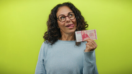 Middle-aged woman shows icelandic krona banknote with playful expression against a vibrant yellow...