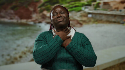 Woman on seaside struggling with neck pain wearing green sweater expressing discomfort amid scenic...