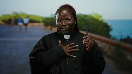 Woman in priest attire holds cross at seaside promenade, evoking spirituality and peace against...