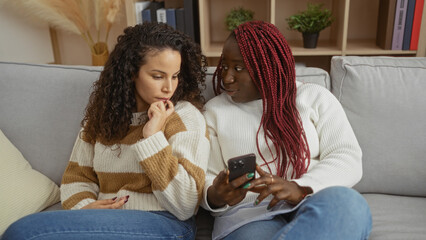 Two women friends sitting indoors on a cozy sofa in a modern apartment discussing something intriguing while one holds a smartphone and the other looks curious and engaged