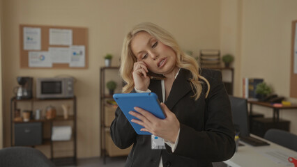 Woman multitasking in modern office talking on phone while using tablet in professional environment...