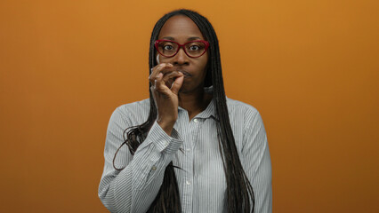 Woman wearing glasses making silence gestures against an orange wall, indicating secretive mood and expression, with a striped shirt in an isolated environment.