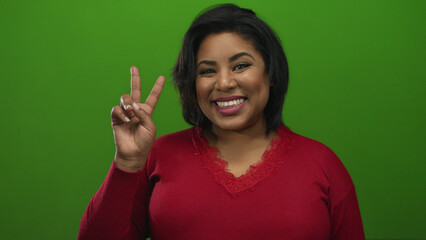 Woman smiling in red sweater showing peace sign against green background expressing joy and...