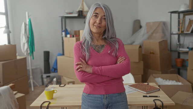 Senior woman with grey hair stands confidently in new apartment living room surrounded by moving boxes, wearing a pink sweater, expressing empowerment and relocation.