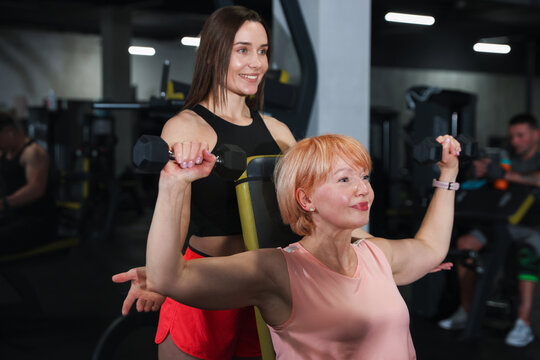 Senior woman training with dumbbells supervised by professional trainer in gym