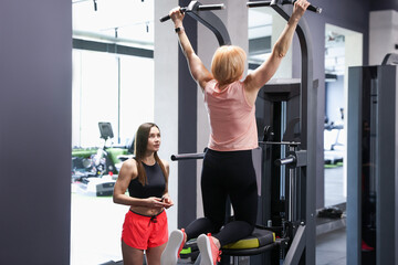 Senior woman training on power tower supervised by professional trainer in gym